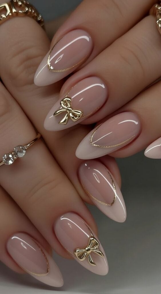 Nude almond nails with fine gold outline and small gold bow charms.