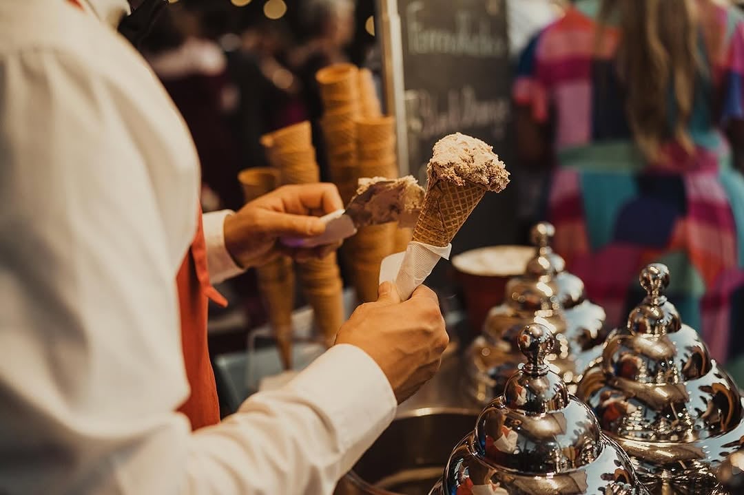 Server holding an ice cream cone beside a gelato cart with stacked waffle cones and metal topping containers in warm low-light candid reception atmosphere