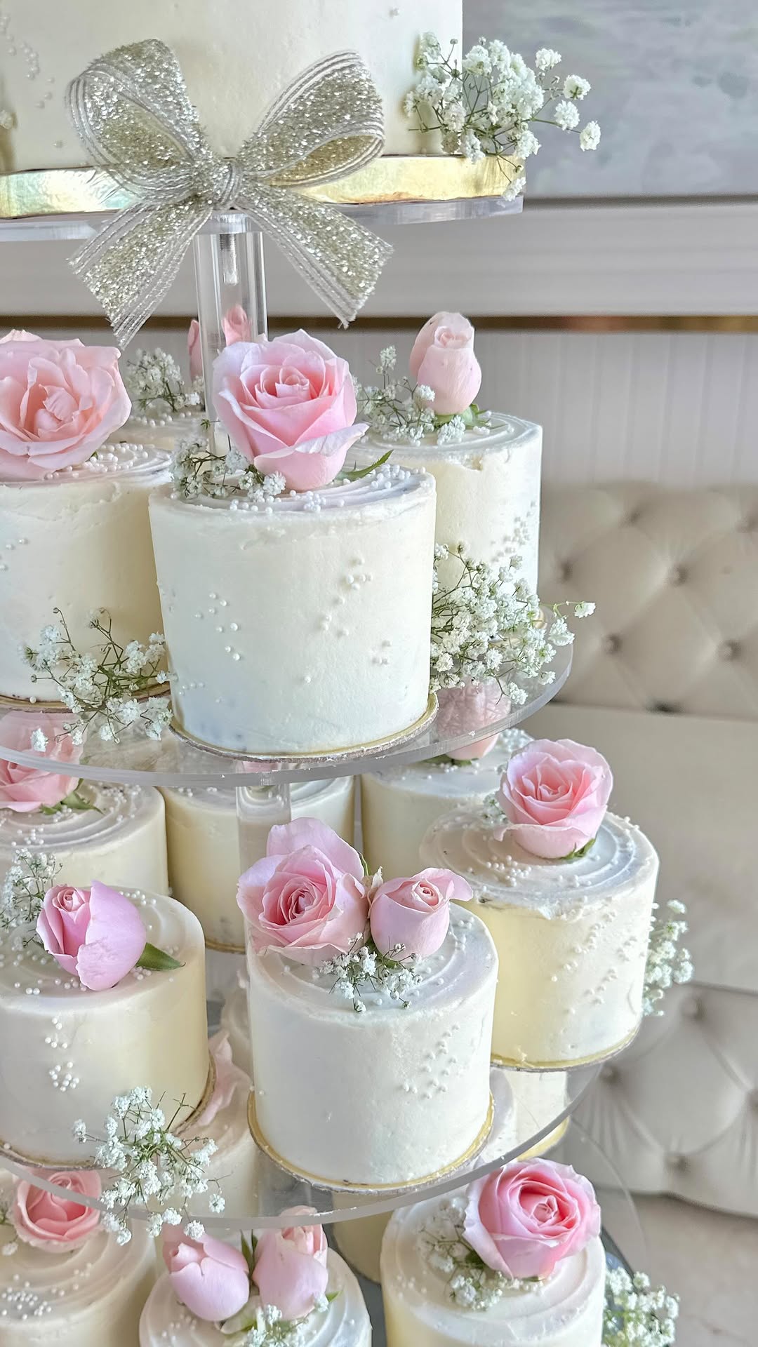 Tiered display of mini cakes decorated with pale pink roses, baby's breath, and pearl detailing in white and blush palette against a refined bridal background