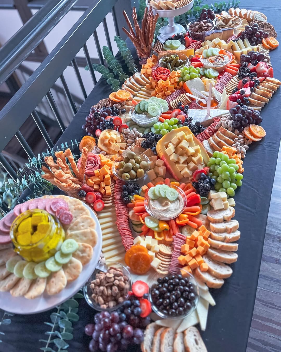 A long dark table packed with an abundant grazing layout featuring cheeses, crackers, fruit, olives, nuts, and dips arranged in circular and crescent shapes with rustic boards and bowls, and a bright yellow dip in the foreground.