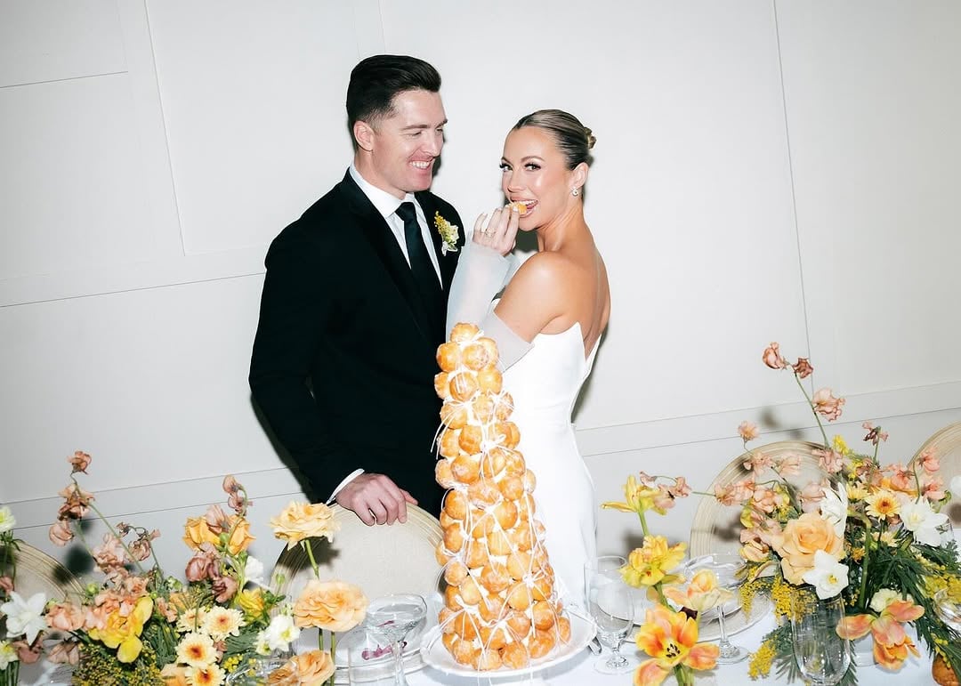 Bride and groom smiling behind a tall donut hole tower with soft yellow and peach floral arrangements in a bright white editorial wedding setting