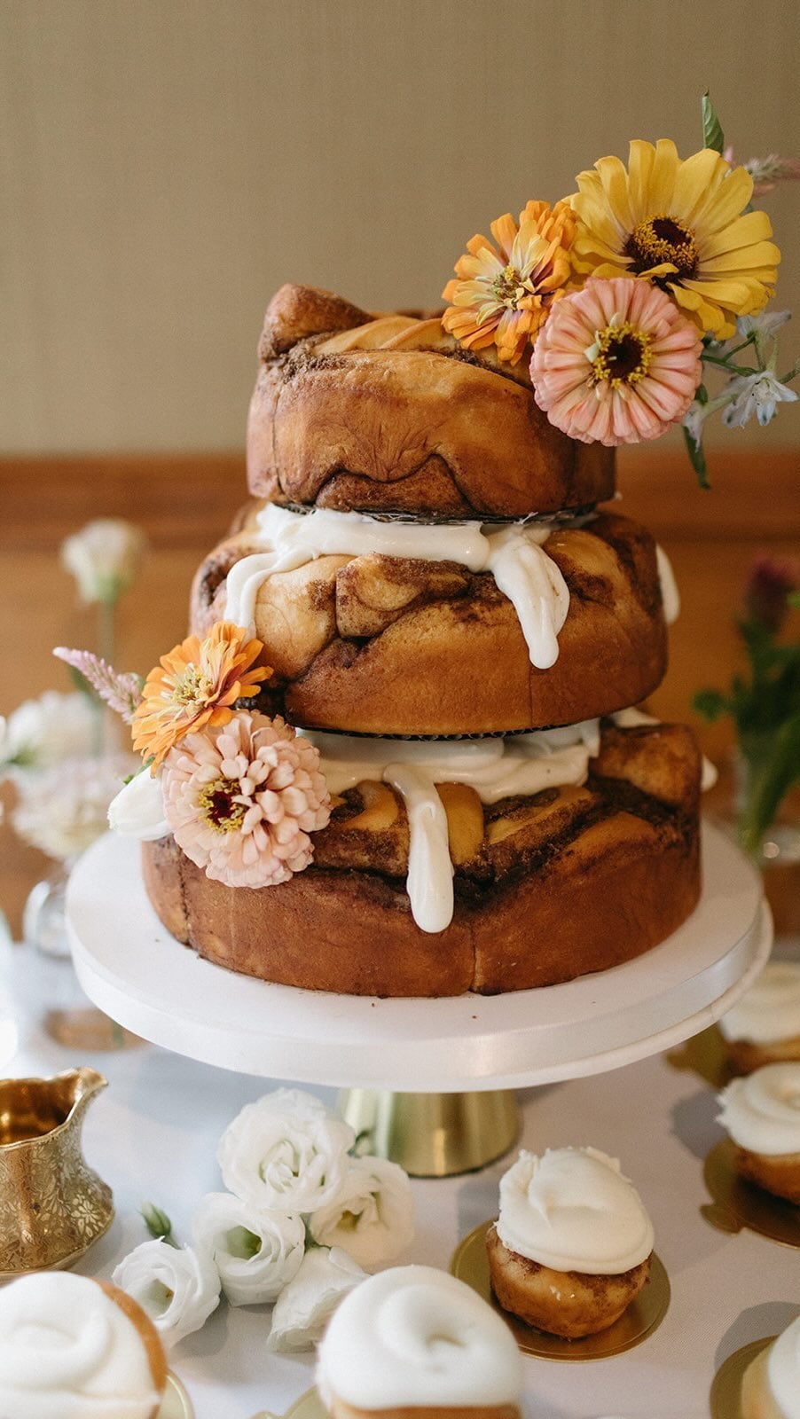 Three-tier cinnamon roll cake on a white stand with thick white icing and warm-toned daisies, set against a moody brown and gold background
