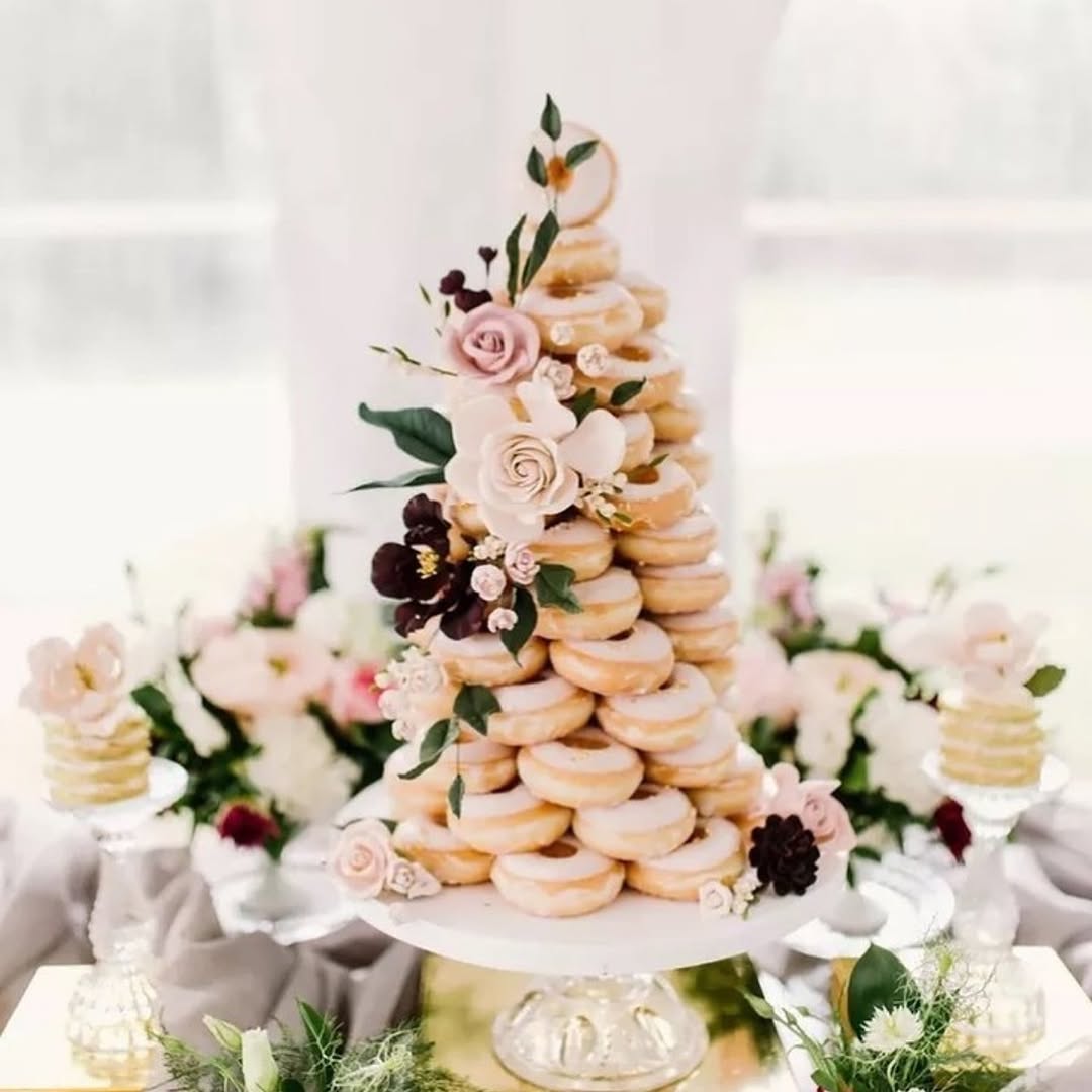 Tiered donut tower resembling a wedding cake with powdered donuts, blush rose floral topper, and dark berry accents on a clean white background