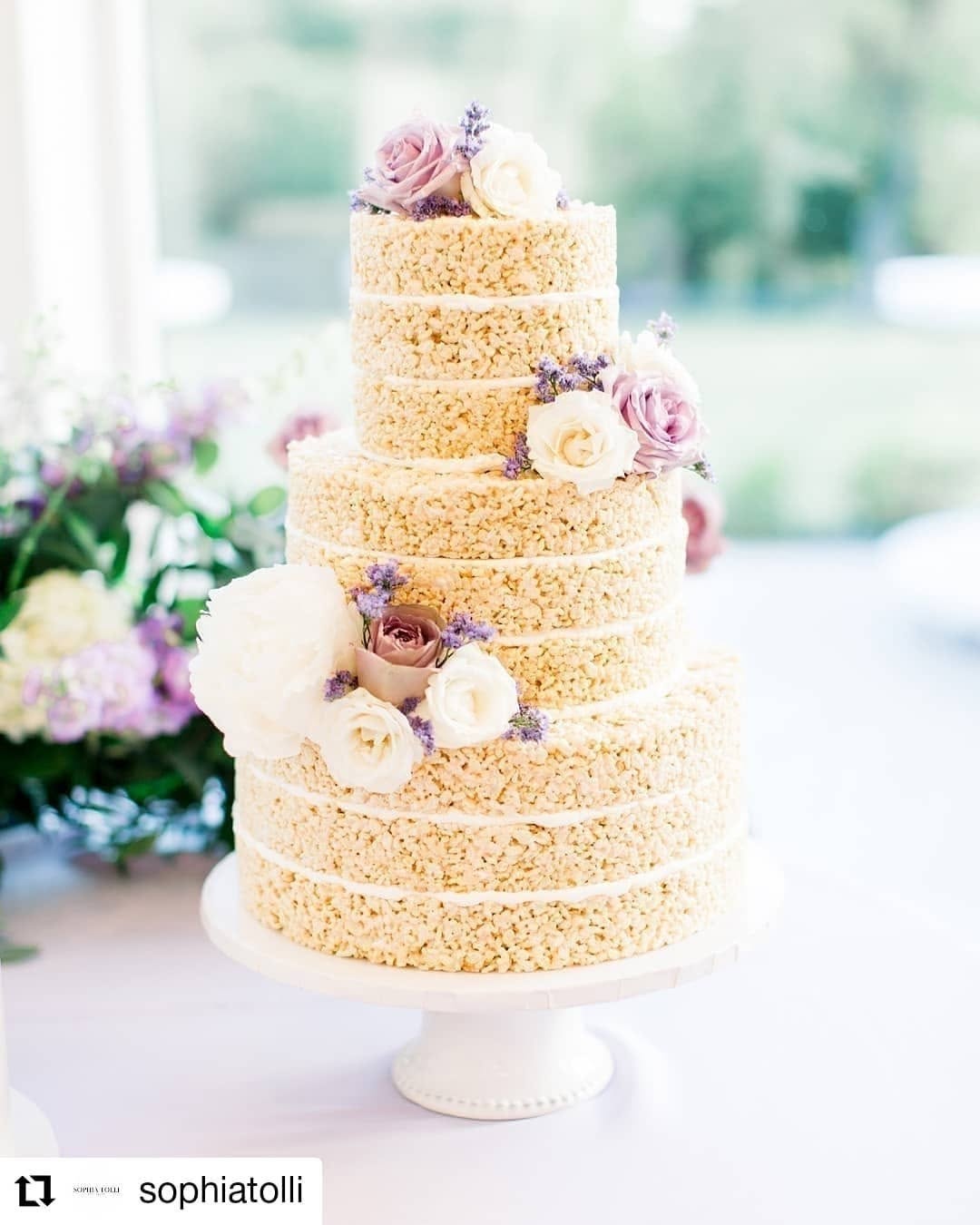Three-tier Rice Krispies treat wedding cake styled as a naked cake with blush roses and greenery between tiers on a white pedestal