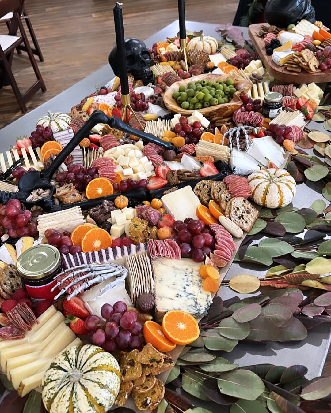 A large grazing table styled for a festive fall theme, with cheeses, meats, fruit, crackers, and greenery garland across the center, small decorative pumpkins tucked among the food, and a rich palette of deep purples, reds, oranges, and warm neutrals.