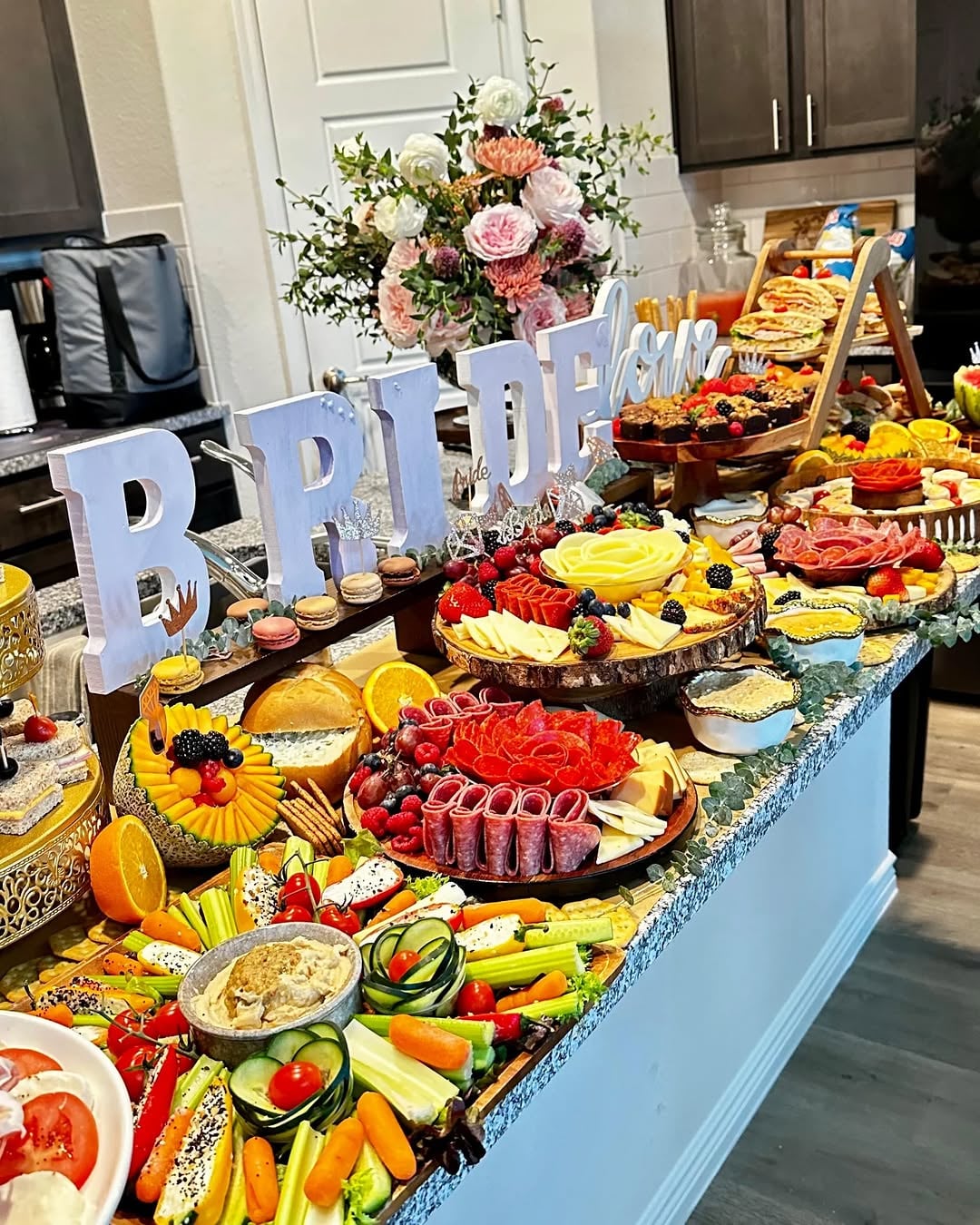 A long kitchen island covered edge-to-edge with a lavish grazing spread featuring sliced cheeses, cured meats, crackers, dips, and colorful fruit clusters, with large block letters spelling BRIDE as the centerpiece and a pastel floral arrangement behind them.