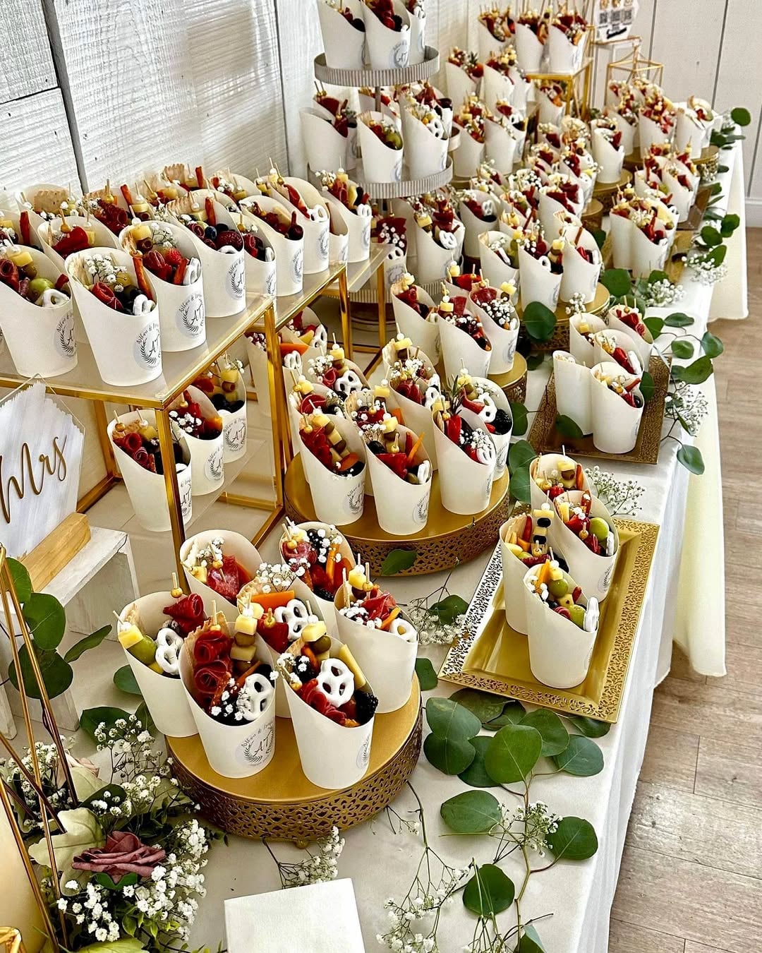 Dozens of individual charcuterie cups arranged tightly on tiered gold risers and trays, each filled with crackers, breadsticks, fruit, cheese cubes, and cured meats, styled on a light tabletop with trailing greenery.