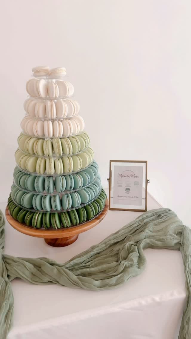 Macaron tower with ombre color gradient from white at the top through pale greens to deep teal at the base, on a wooden board with green draped fabric