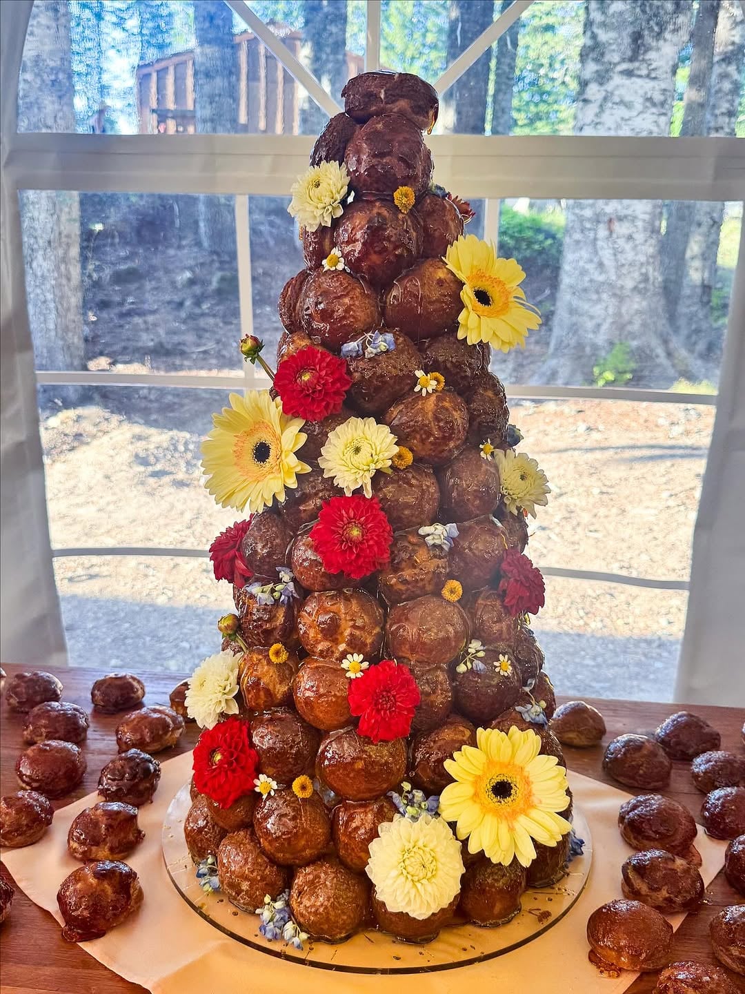Croquembouche tower of caramel-glazed cream puffs decorated with yellow, red, and white fresh flowers on a wooden table with forest view