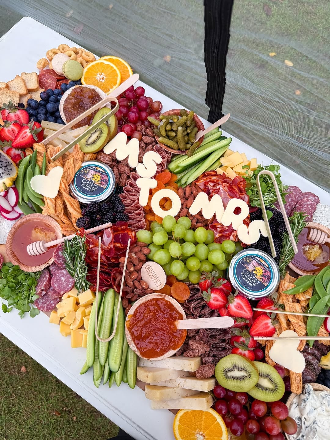 An overhead view of an outdoor grazing display featuring a MRS TO MR sign nestled among cured meats, cheeses, grapes, strawberries, pickles, and dips on a gray tabletop with bright natural light.