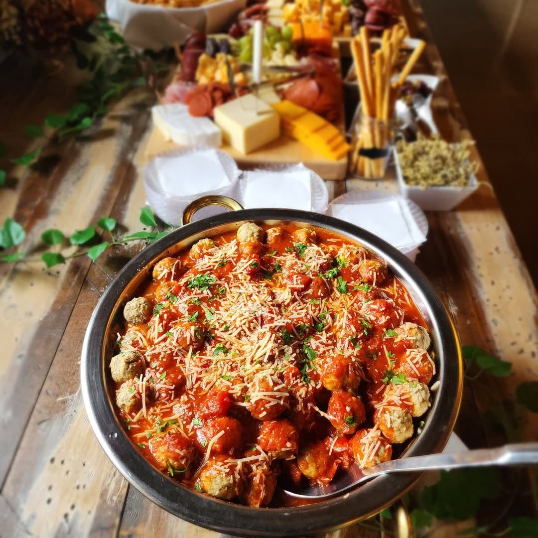 A close-up of a baked appetizer in a small cast iron skillet topped with herbs and cheese, set on a rustic wooden board with charcuterie elements including cheeses, crackers, cured meats, and small bowls visible behind it.