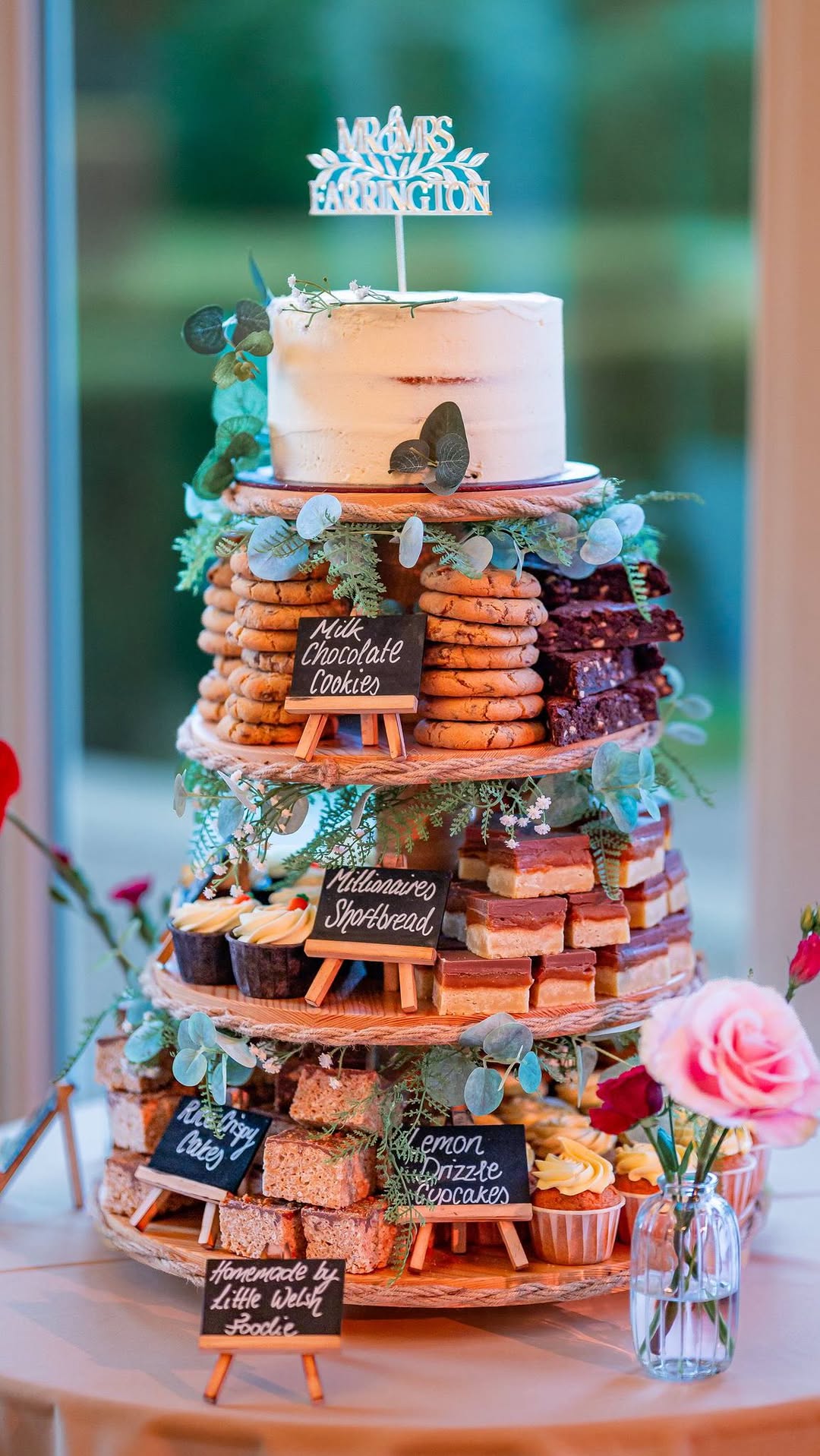 Tall wedding cake tower built from layered trays of assorted baked bars and treats with chalkboard labels, floral topper, and soft indoor lighting