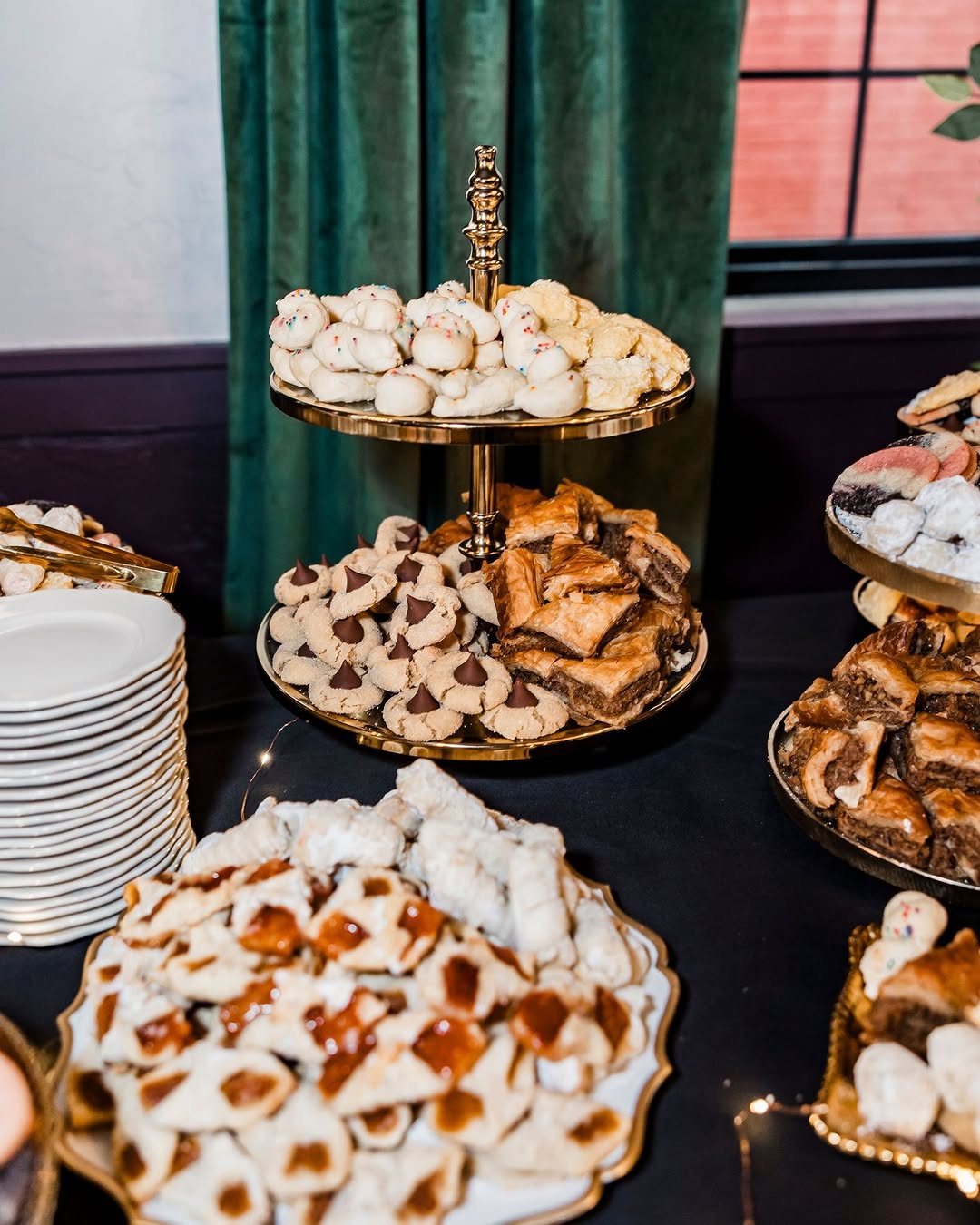 Abundant cookie and pastry dessert station with meringues and layered sweets on gold stands against dark green drapery in a moody luxe reception setting