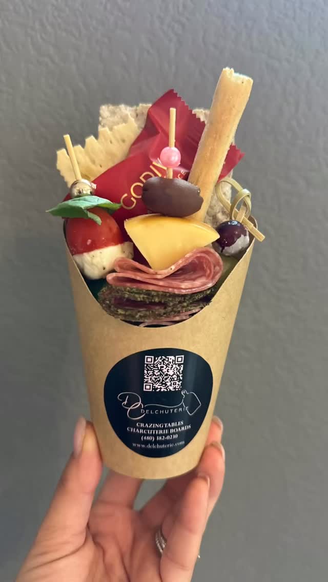 A single charcuterie cone held in one hand against a plain gray background, filled vertically with breadsticks, folded cured meats, cheese, grapes, berries, and a strawberry, with a circular branded label and QR code visible on the cone.