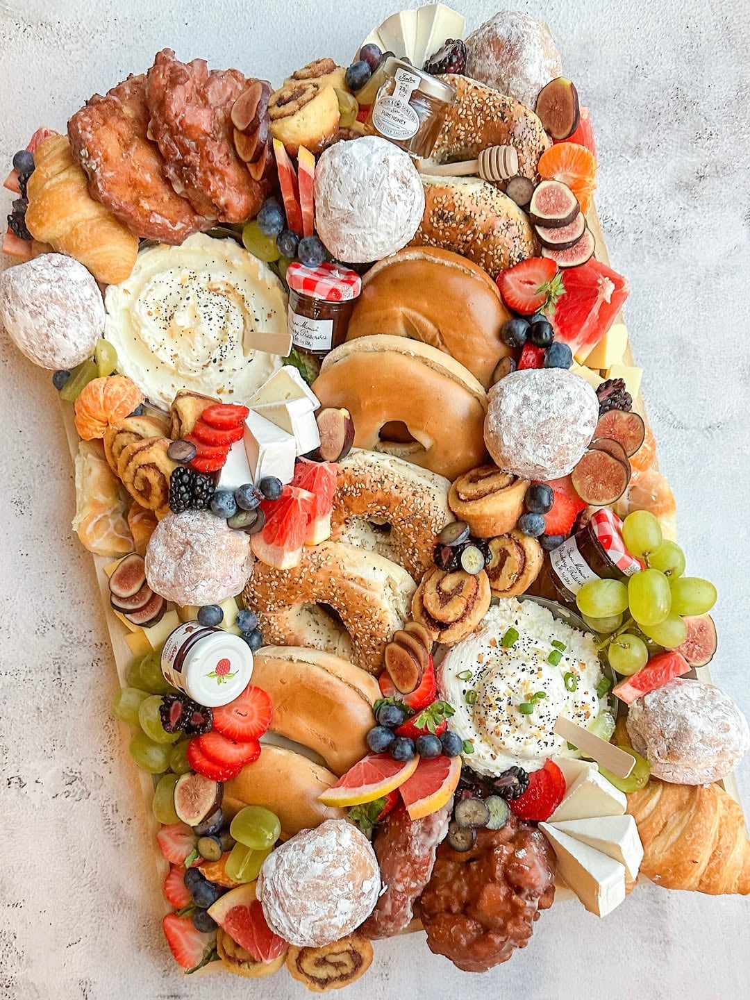 A top-down view of a brunch-style grazing board loaded with bagels, pastries, fruit, spreads, and assorted bites in light and fresh colors of creams, tans, and berry reds, spread densely across a pale surface with small bowls breaking up the texture.