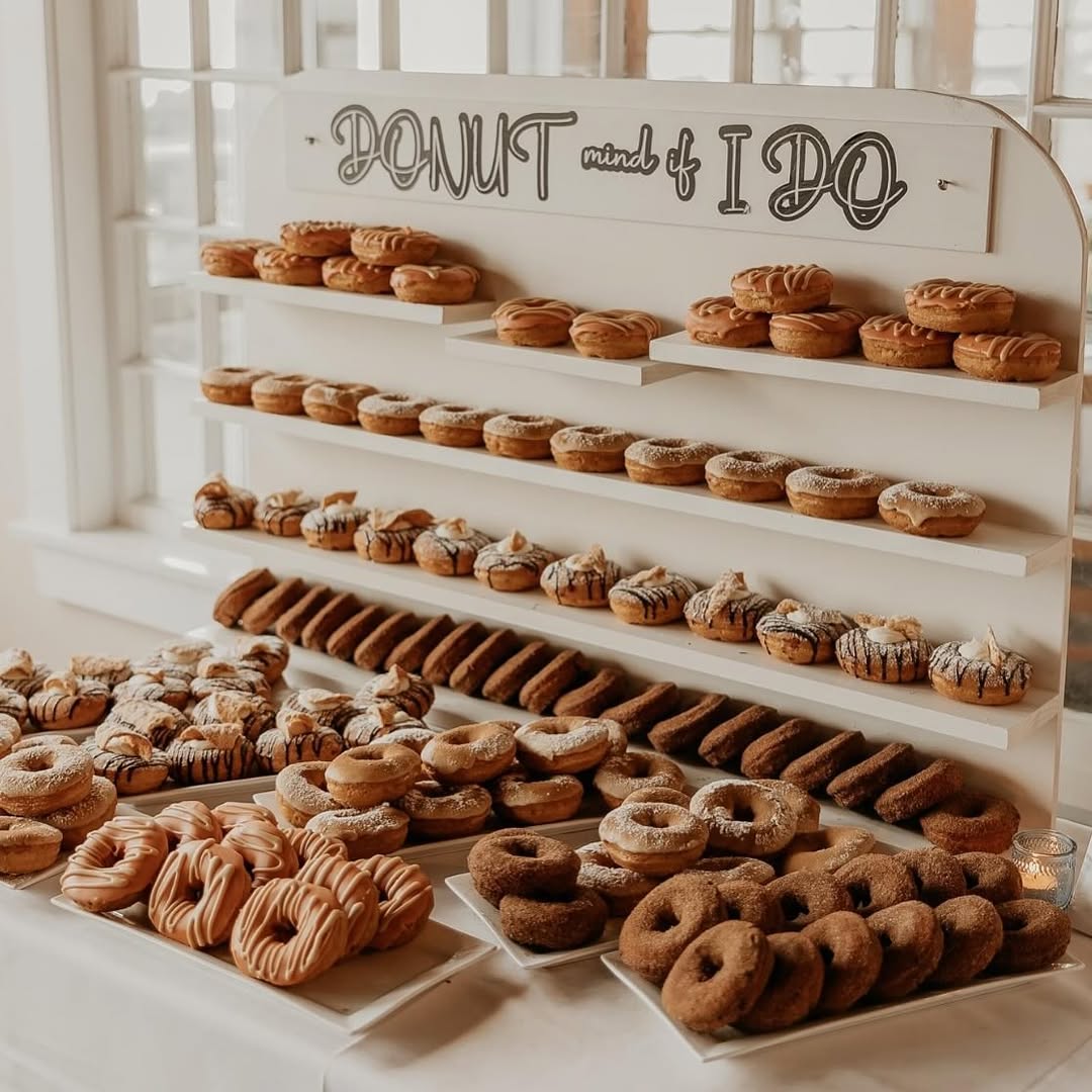 Donut wall with rows of donuts in a grid pattern as a backdrop, additional pastries on platters flanking the display in clean white and neutral styling