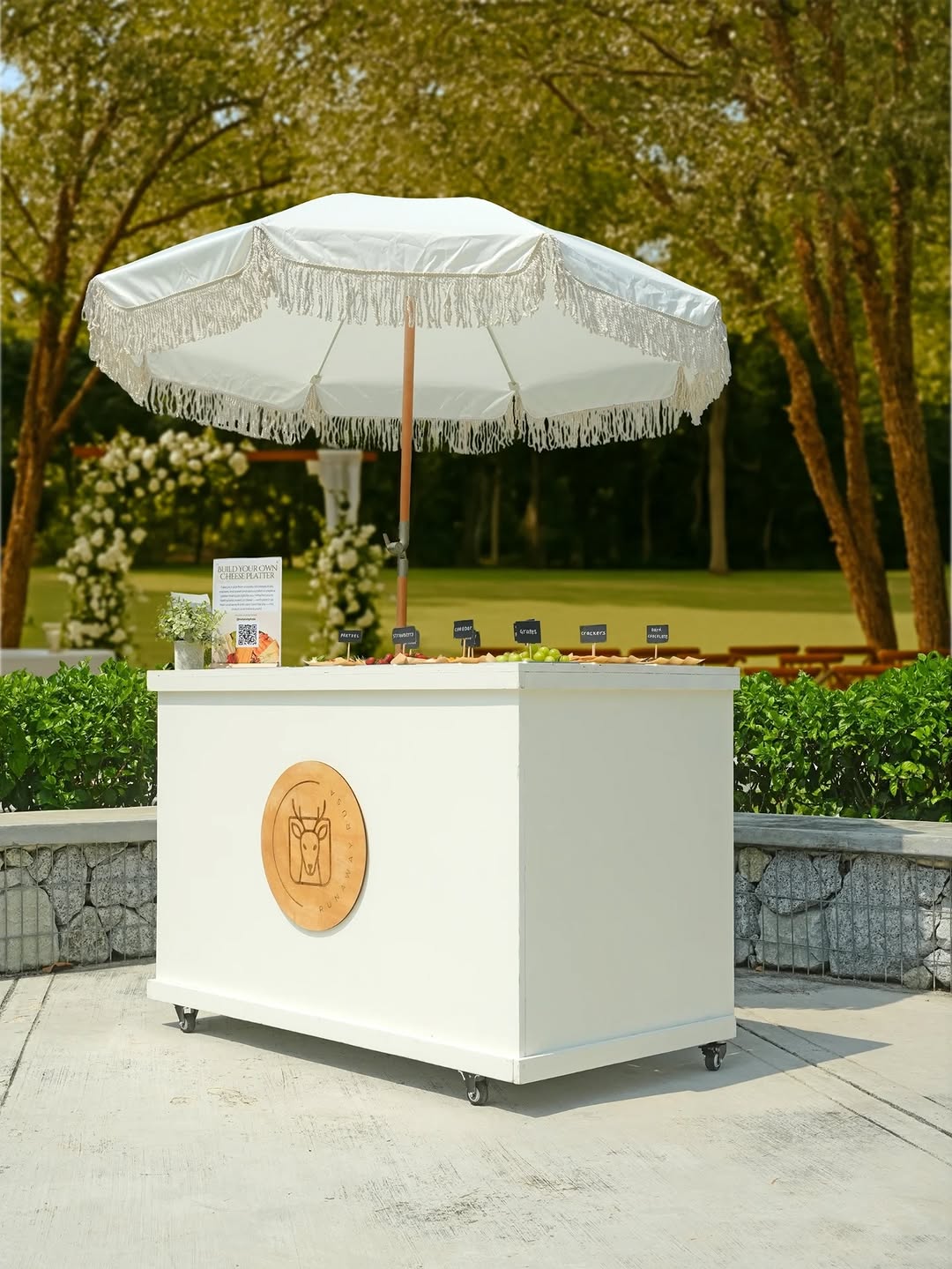 A sleek white branded catering cart set up outdoors on a lawn, topped with neatly arranged individual boxes or trays in a grid-like layout, with rows of wooden ceremony chairs and hedges visible in the background suggesting a wedding venue setting.