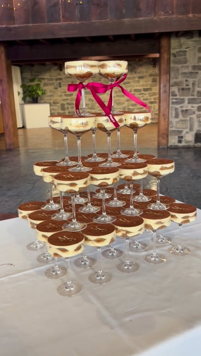 Wide tiered tiramisu tower with cups on wine glass stems, small upper tier tied with red ribbon, in a stone-walled event space with warm reception lighting
