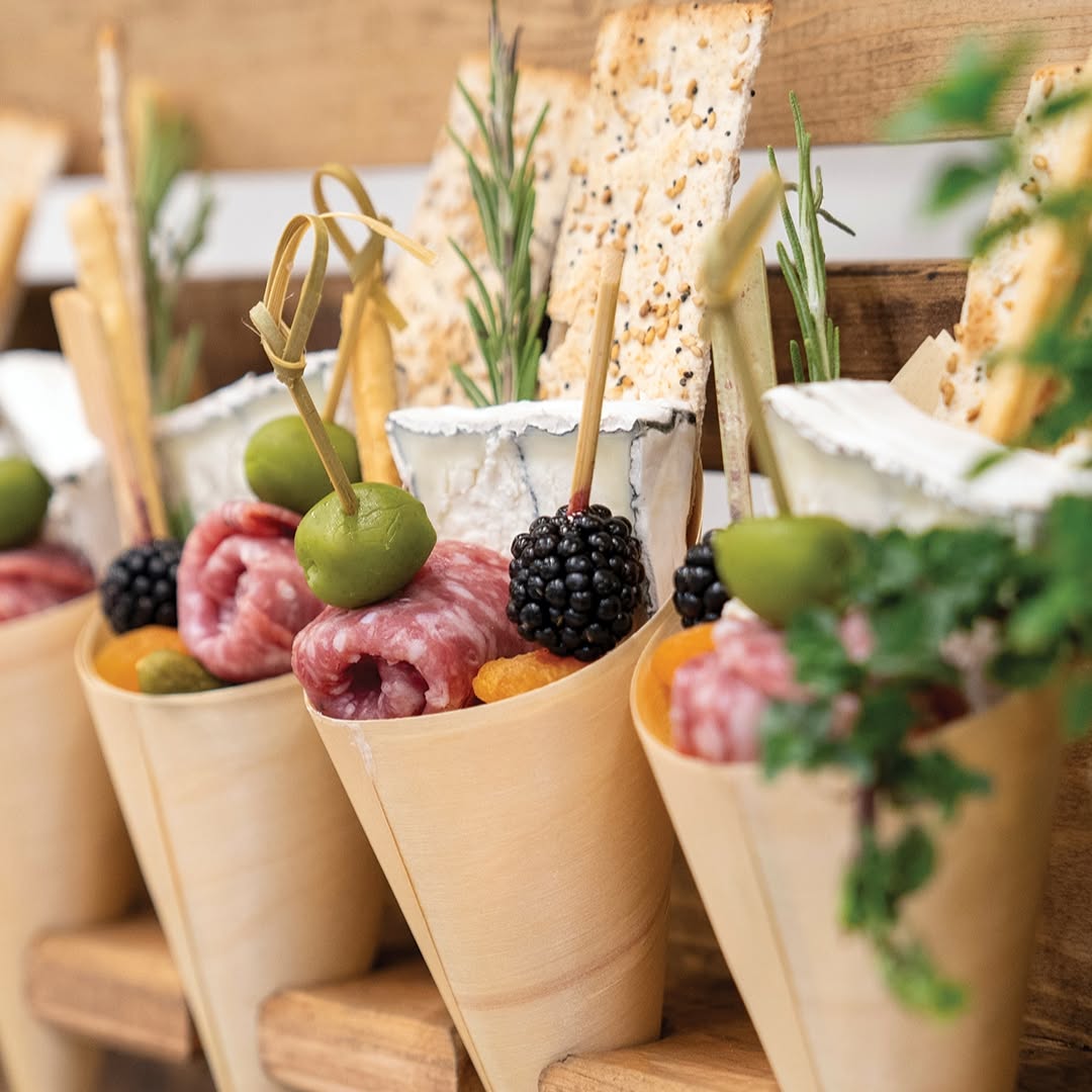 Several charcuterie cones in a row on a wooden surface, filled with breadsticks, cheese wedges, olives, berries, and fresh herb garnishes, with shallow depth of field making front cones crisp while the rest blur into the background.