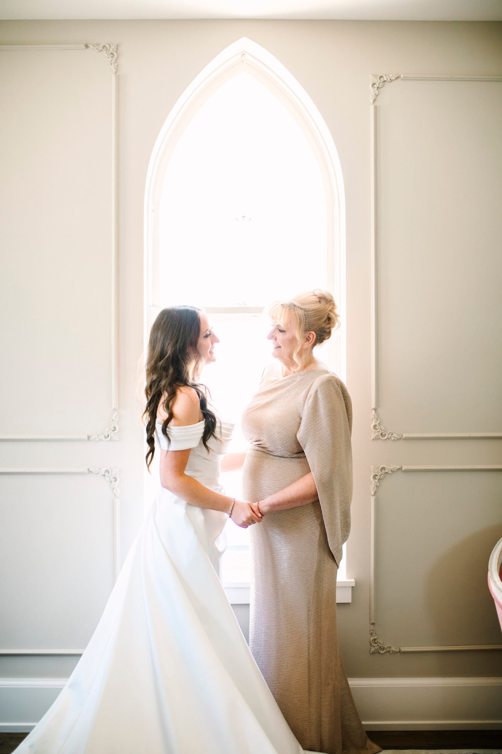 Abigail and her mom holding hands in front of an arched window before the ceremony
