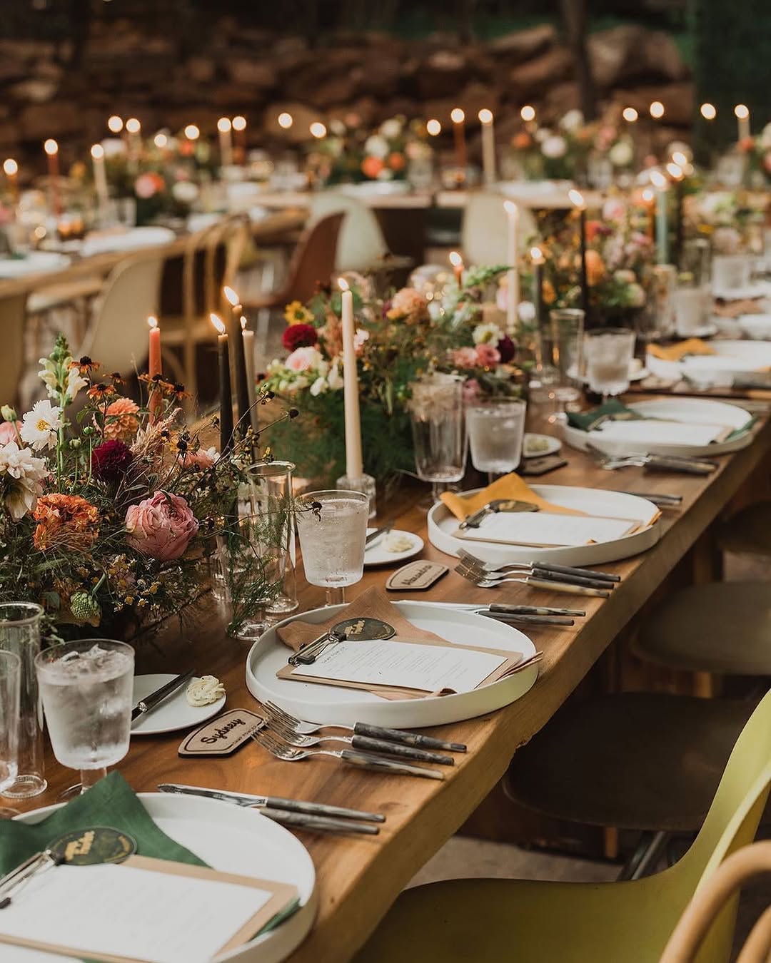 Warmly lit evening reception table with layered place settings, mixed-height taper candles, small muted floral clusters, and soft string lights glowing in the background