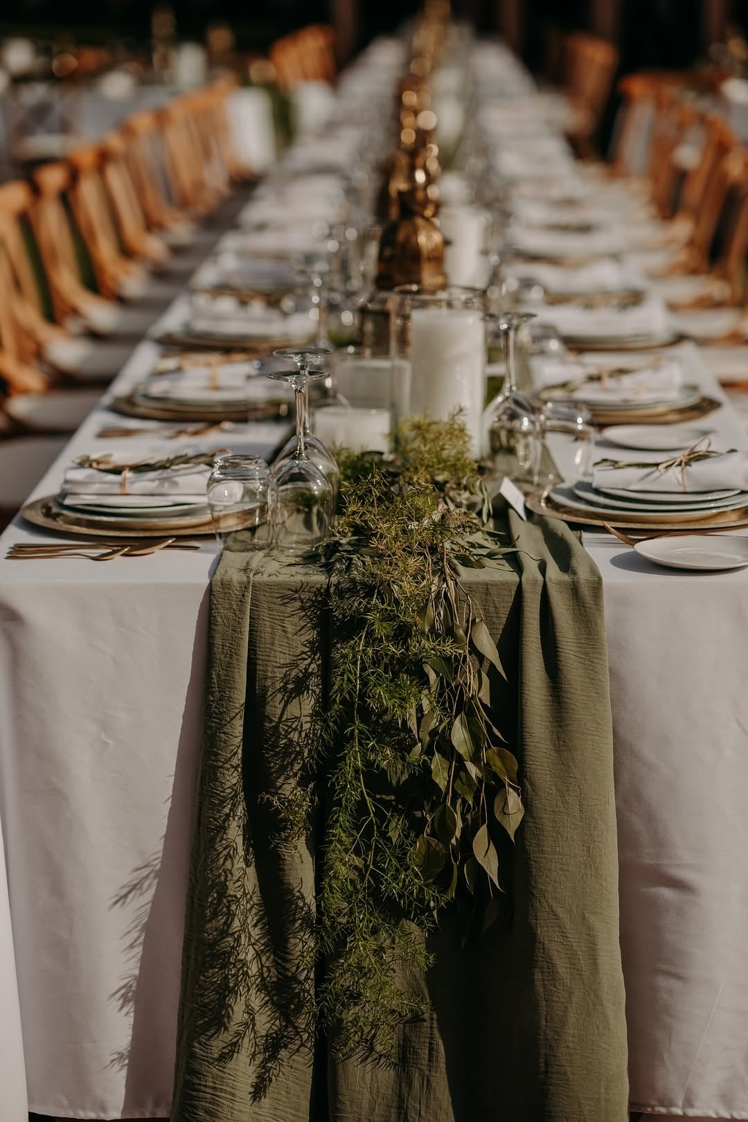 Long outdoor banquet table with sage green draped runner, trailing greenery, and clear glass candle holders with white pillar candles