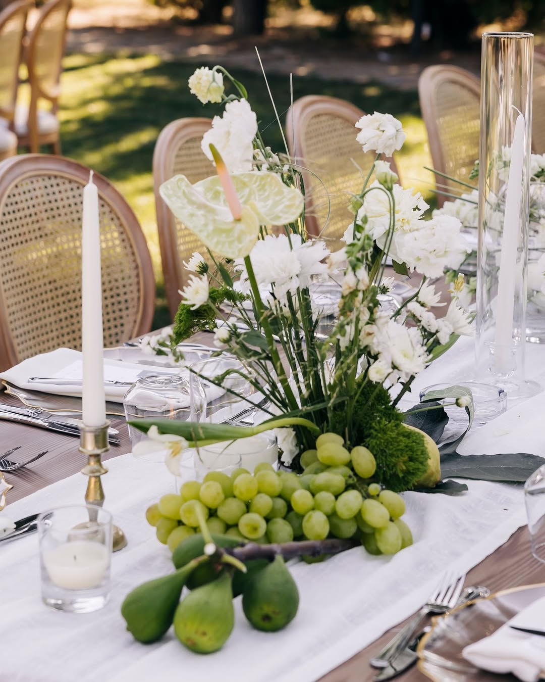 Close-up of a garden table with white floral centerpiece, clusters of green grapes and figs, and slim taper candles in clear holders