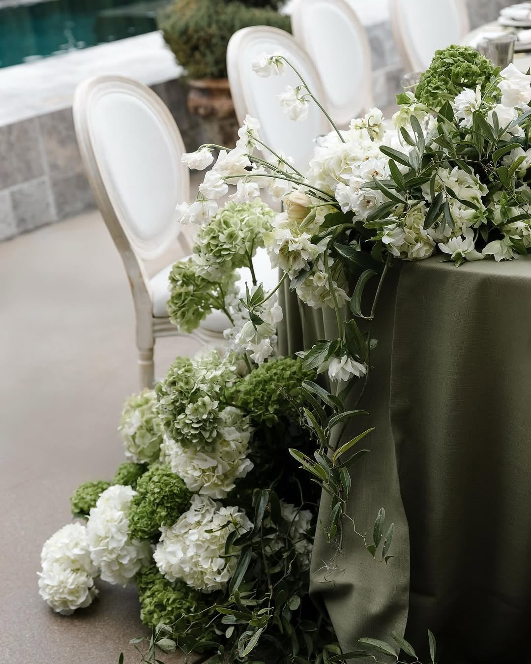 Outdoor reception table with deep olive-green tablecloth, abundant white hydrangea-like florals and greenery spilling over the edge, and rounded white chairs