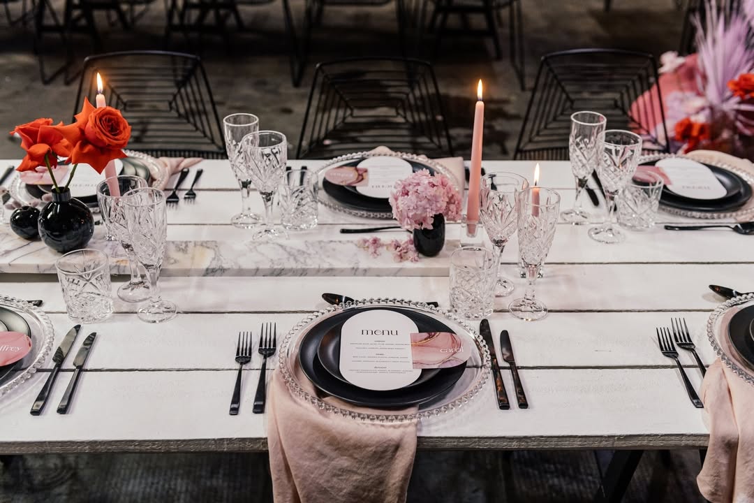 Modern wedding table with a whitewashed wood top, matte black dinner plates on clear beaded chargers, blush pink silk napkins, cut crystal stemware, red roses in small black bud vases, and slim pink taper candles