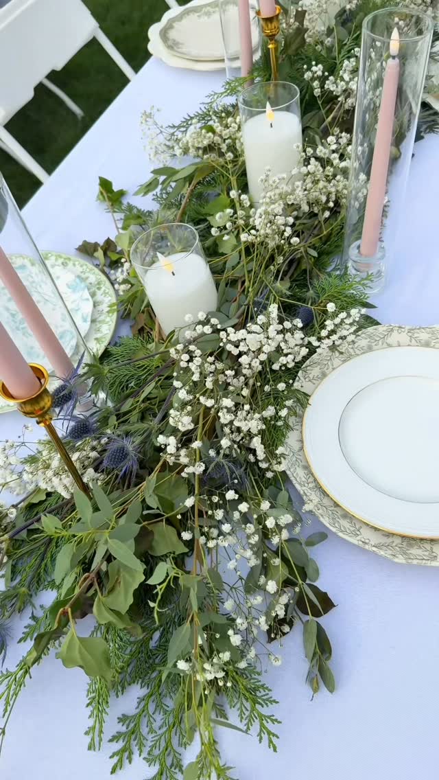 Top-down angled view of a table with pale blue linen, baby's breath greenery runner, white pillar candles, blush taper candles, and gold-rimmed white plates