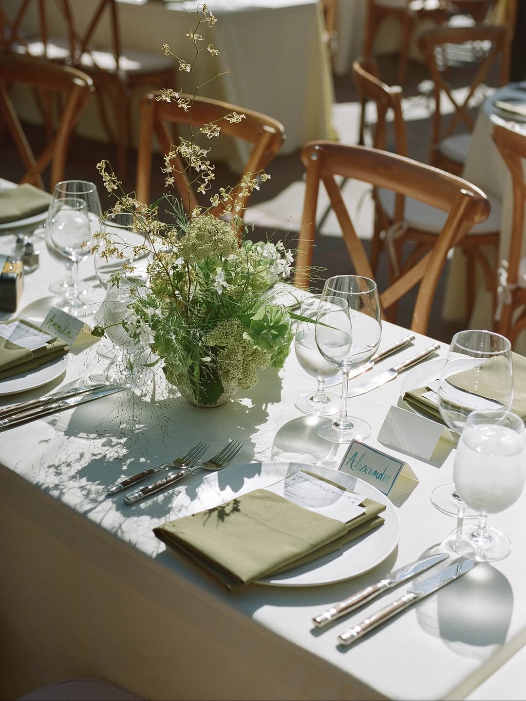 Sunlit outdoor wedding table with crisp white linen, pale sage folded napkins on white plates, clear stemware, wood cross-back chairs, and a compact green and white wildflower centerpiece in a glass vessel