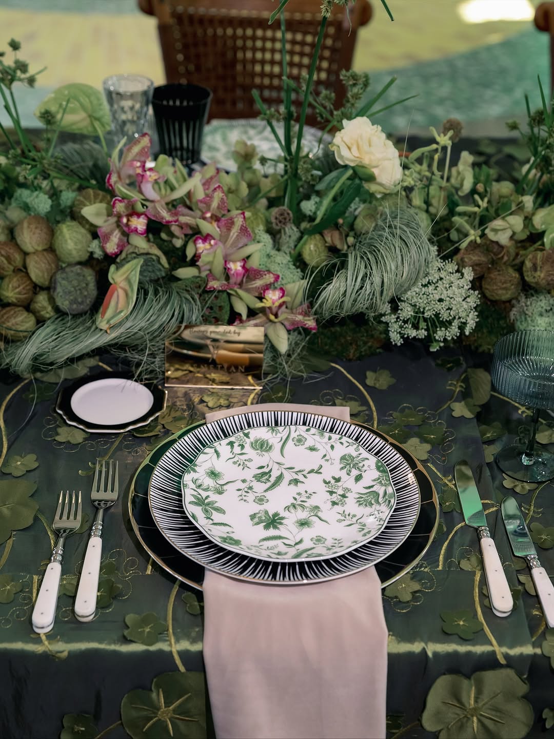 Moody wedding tablescape with a deep green tablecloth, green botanical print dinner plates on black striped chargers, a lush pink orchid centerpiece with green pods and trailing stems, and a blush silk napkin