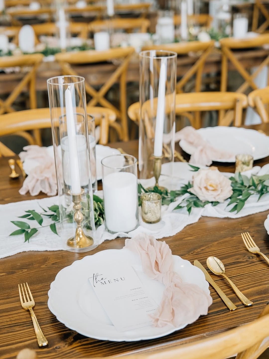 Summer reception table with blush ruffled napkins, clear glass cylinder candle holders, gold flatware, white dinner plates, and a blush rose and greenery runner on a wood table