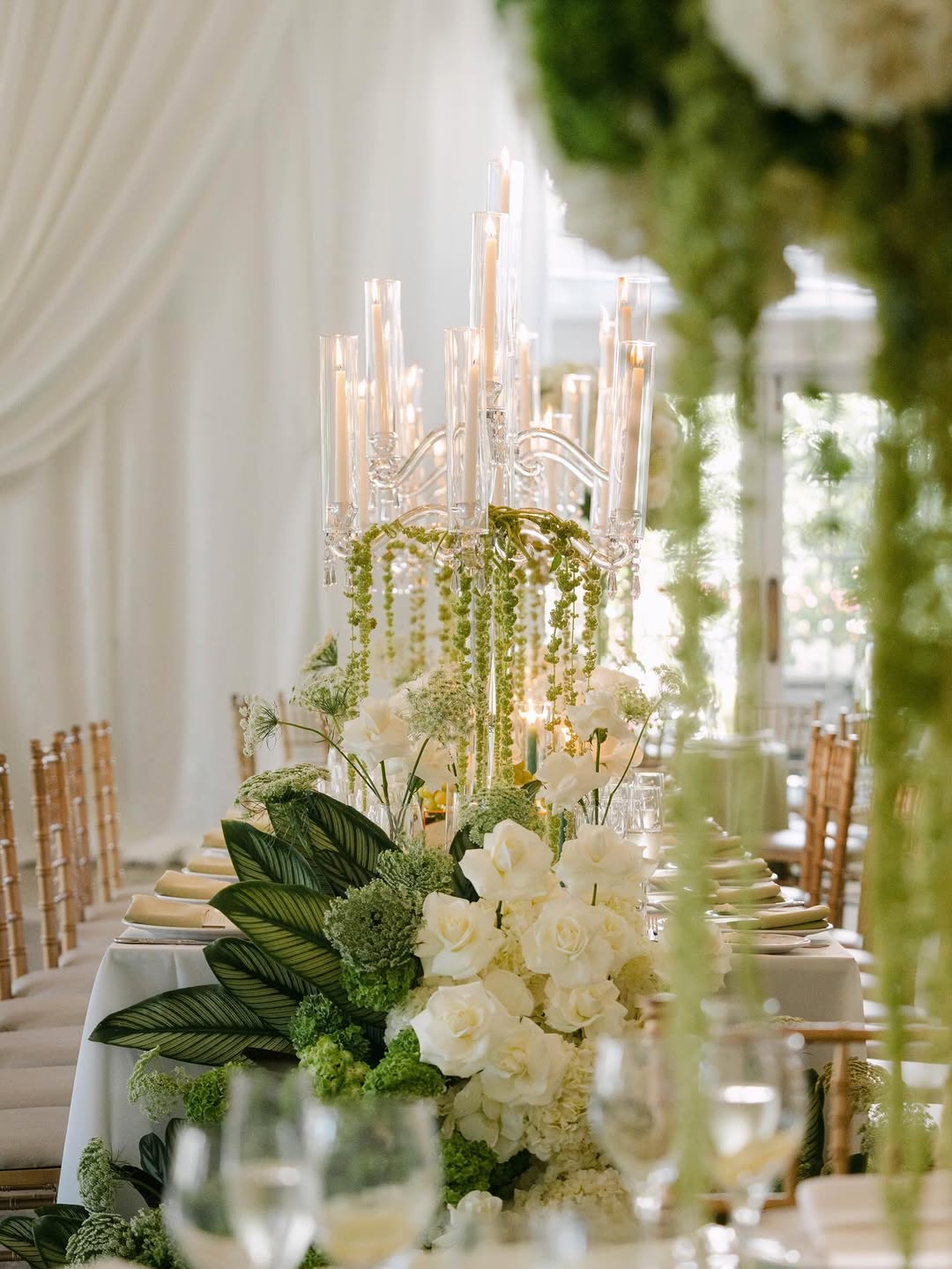 Indoor long reception table with tall crystal candelabra, cascading white florals and greenery, and gold chairs under soft fabric drapery