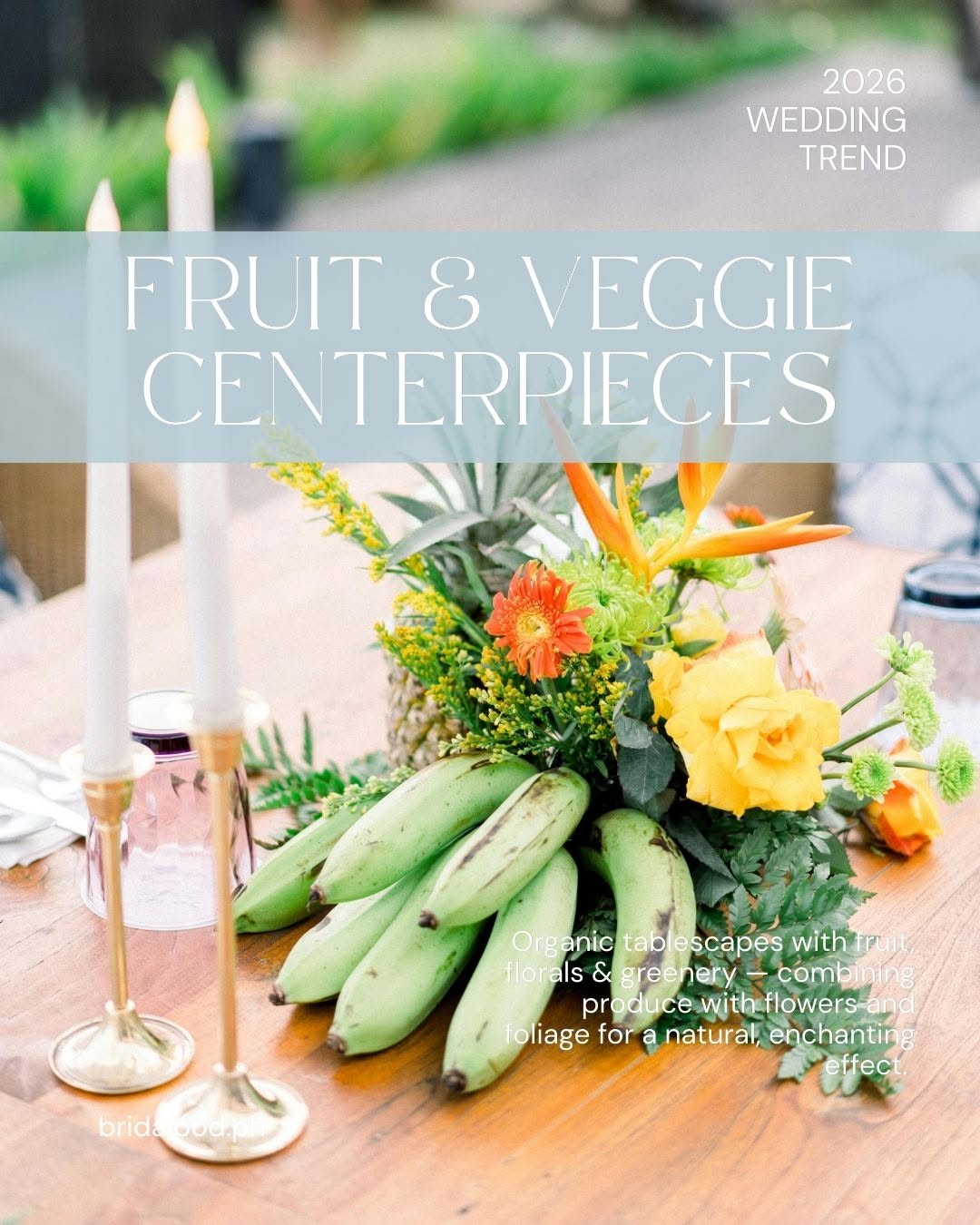 Wood wedding table with a centerpiece of green bananas, yellow and orange garden roses, protea, and taper candles in brass holders next to a small pink bottle