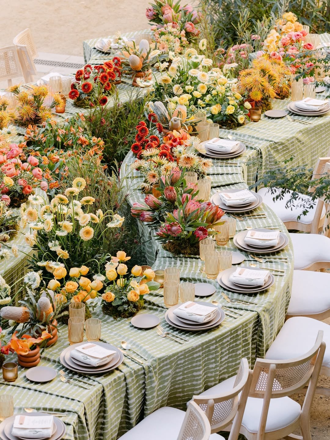 Vibrant tablescape with a patterned green tablecloth, multiple small colorful wildflower arrangements in warm tones, taper candles, and white chairs