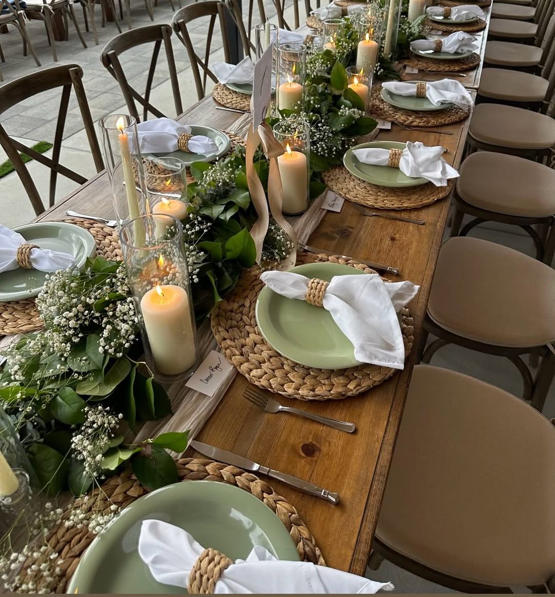 Rustic wood table with green plates, woven chargers, white napkins, and a lush greenery garland with small white blooms and glass votive candles