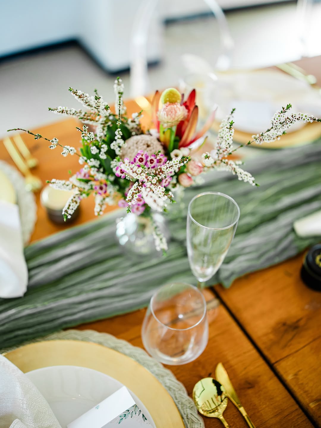 Overhead view of a wood table with a gauzy sage green fabric runner, a compact peach and coral wildflower centerpiece, gold flatware, and clear glassware