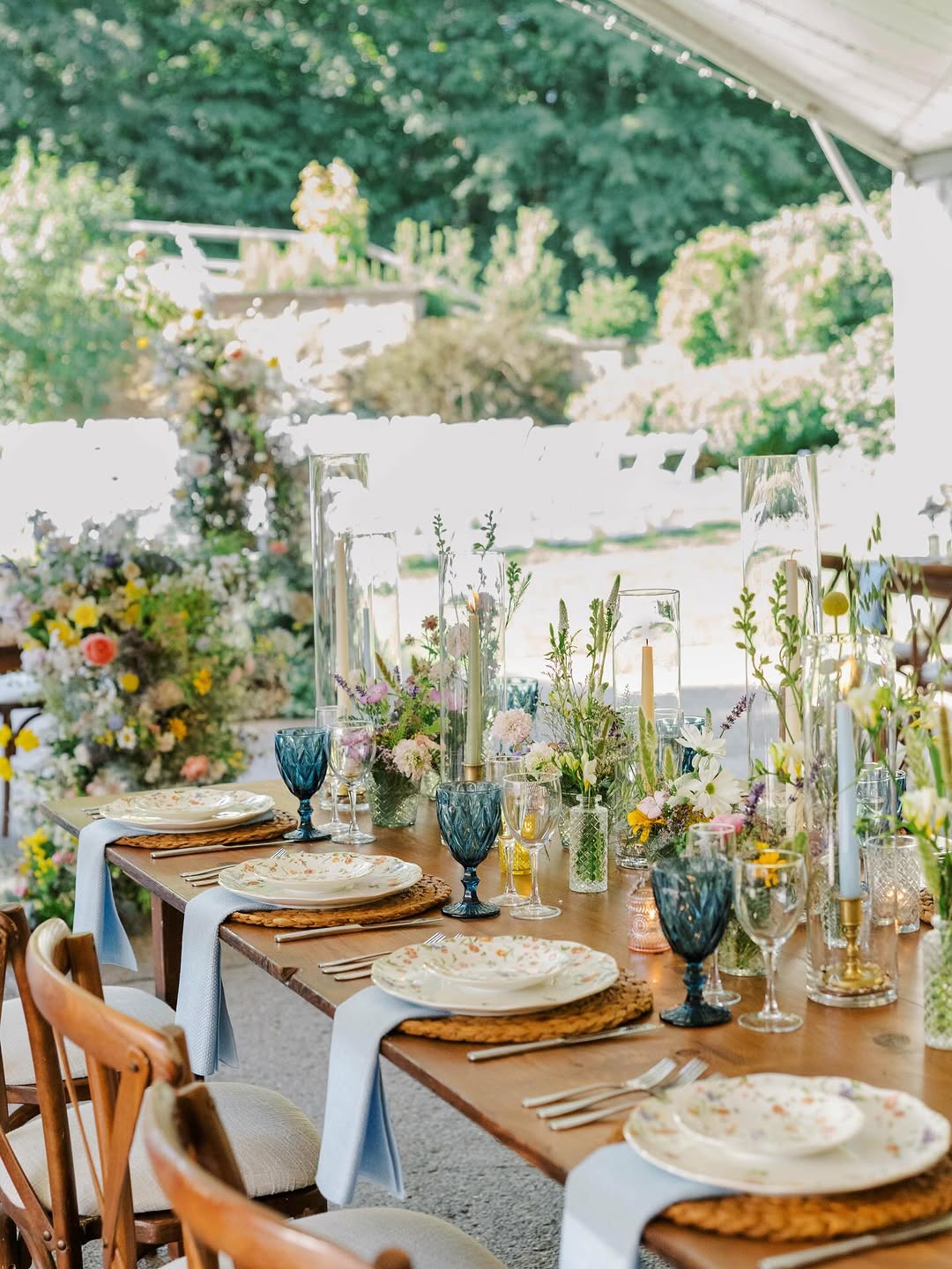 Close-up place setting with a soft blue napkin on a gold-rimmed plate and woven charger, small amber and blue glass bud vases with delicate stems, and clear stemware