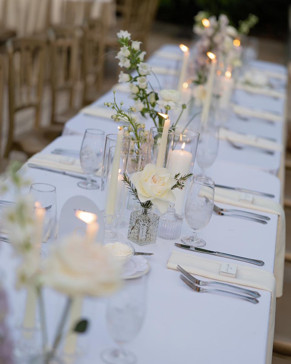Long white linen reception table with a repeating line of small clear bud vases holding single white roses and fresh stems, slim taper candles in clear holders, and natural wood cross-back chairs