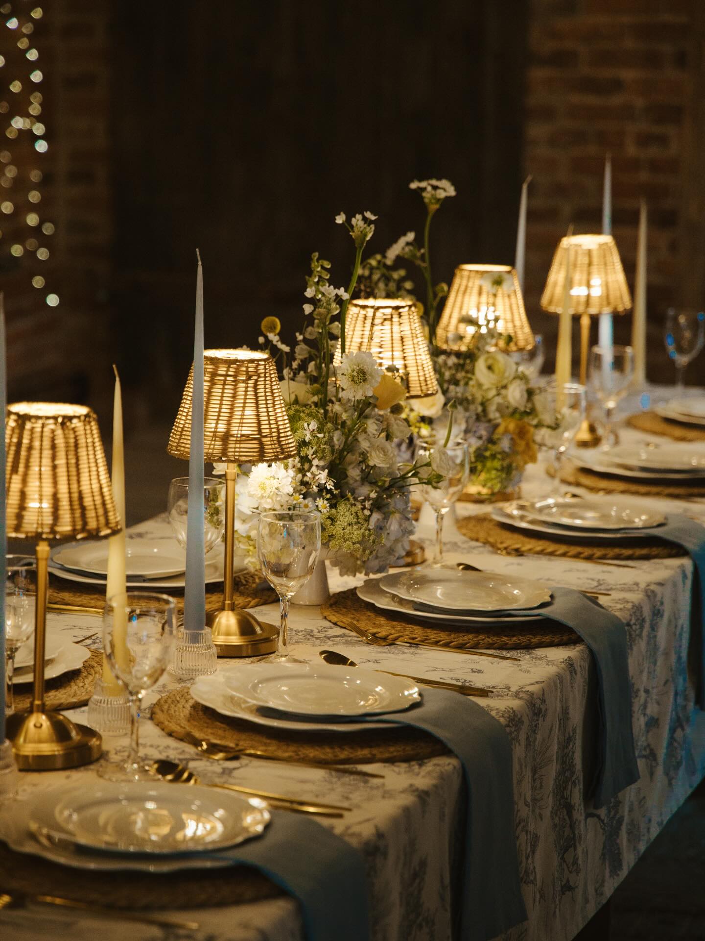 Cordless rattan-shade table lamps on long wedding table with candles and blue toile linen