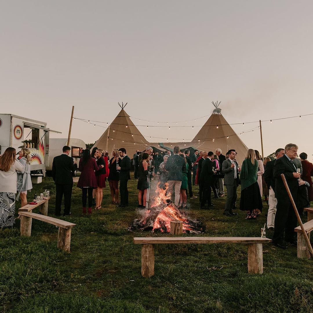 Outdoor wedding bonfire and tipi tents with guests gathered at dusk, Camp Sunny Stones