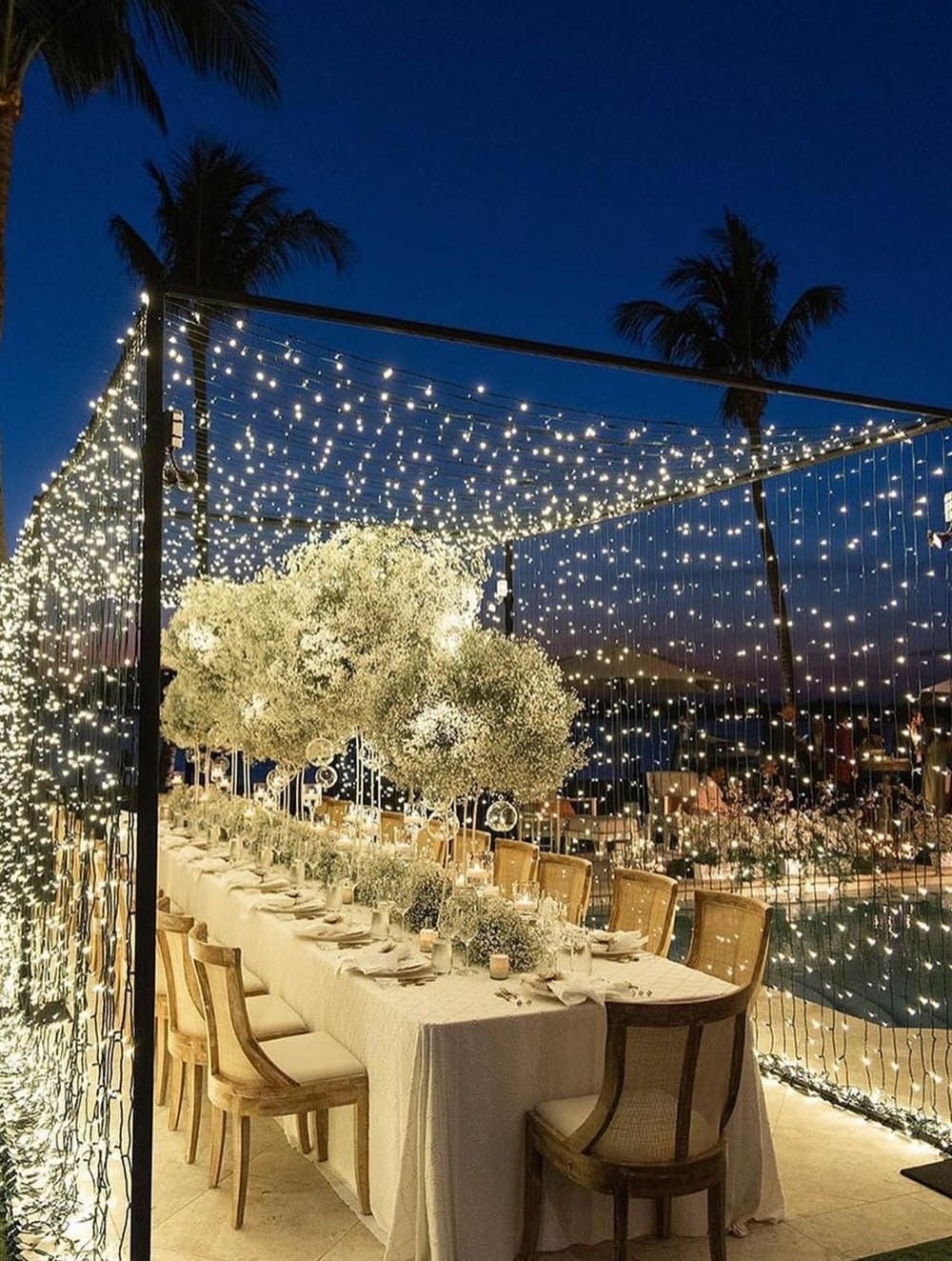 Outdoor pool-side wedding dinner under vertical fairy light curtain at twilight with palm trees