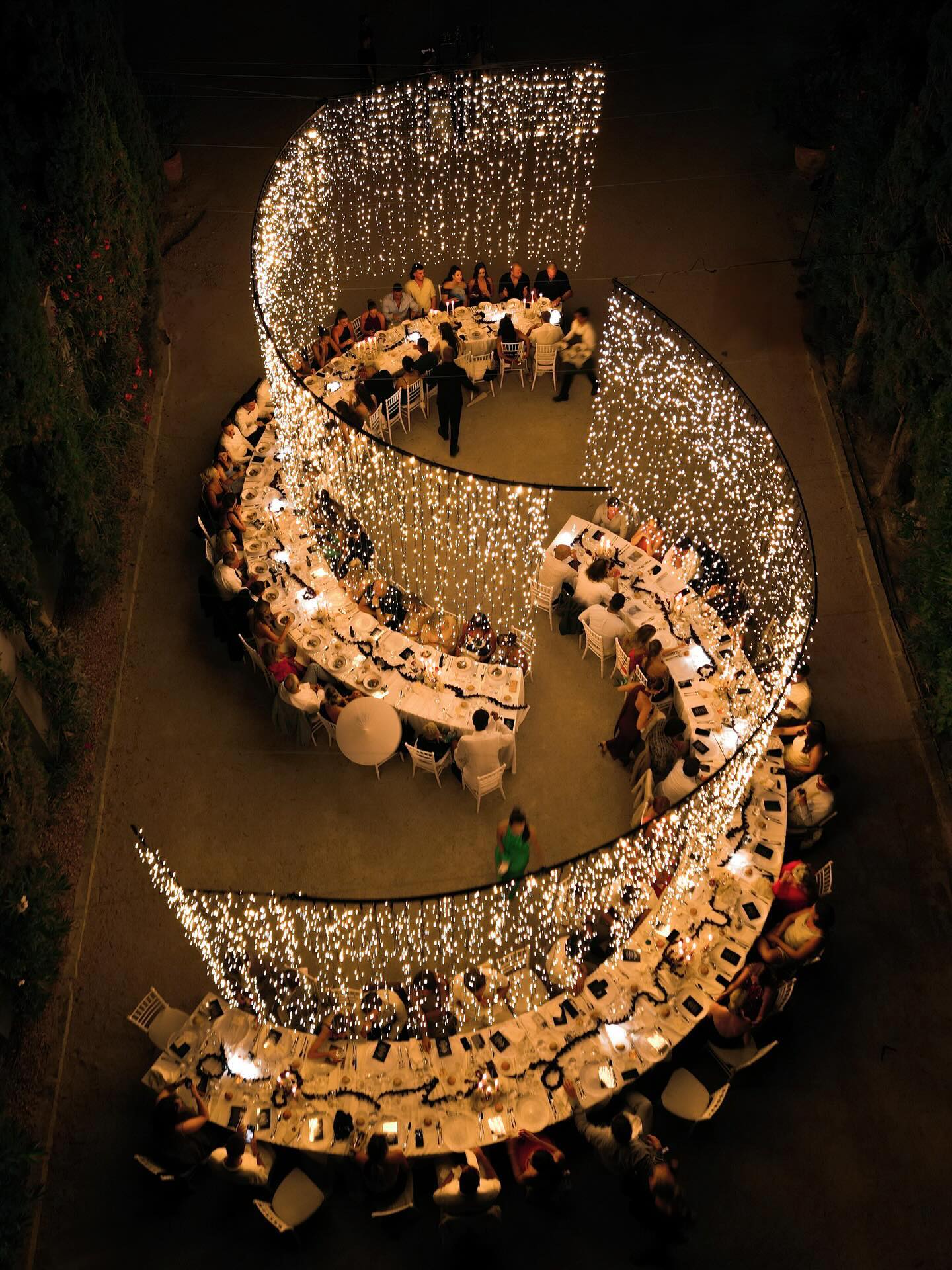 Outdoor wedding reception with S-curved tables under massive cascading fairy light installation, drone view