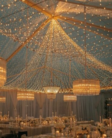 Clear-top tent reception with cascading fairy lights and Edison chandeliers overhead at outdoor evening wedding