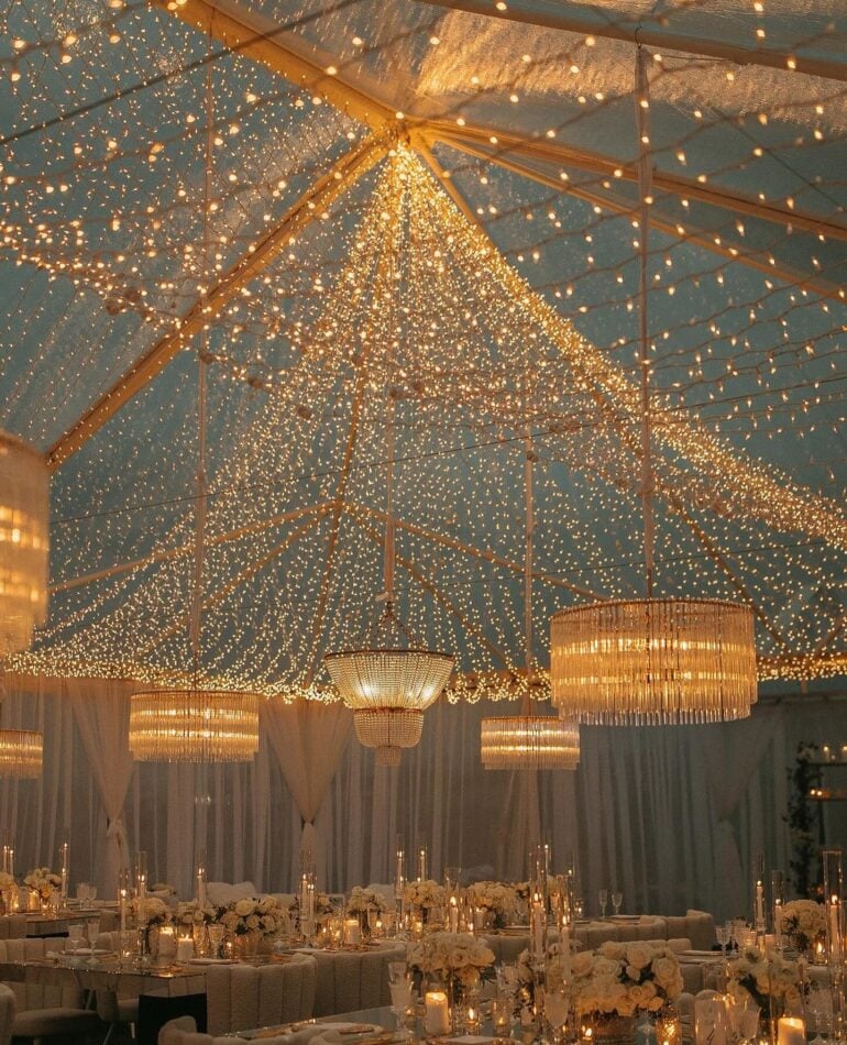 Clear-top tent reception with cascading fairy lights and Edison chandeliers overhead at outdoor evening wedding