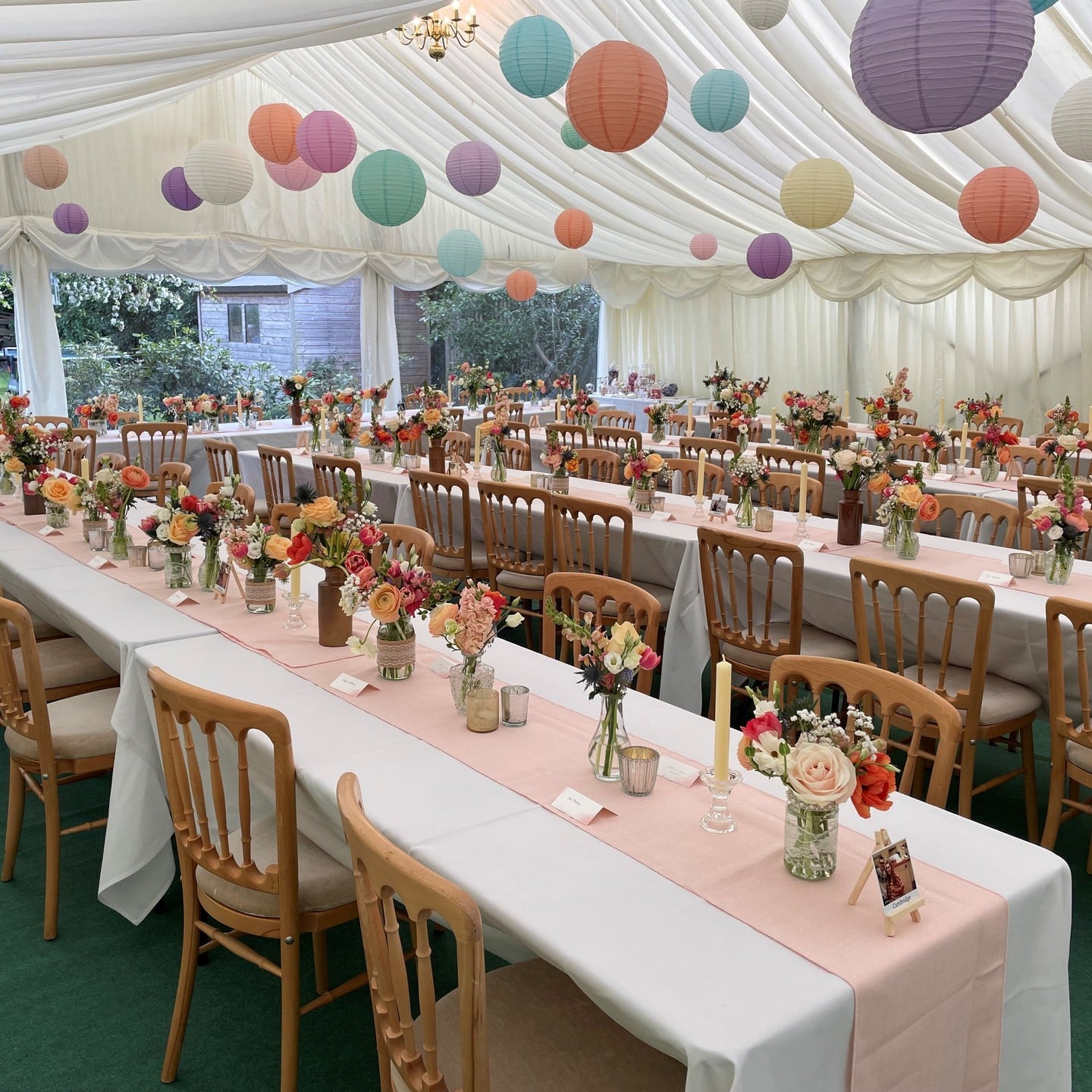 Pastel paper lanterns hanging from white marquee tent ceiling over long farm tables with bright florals