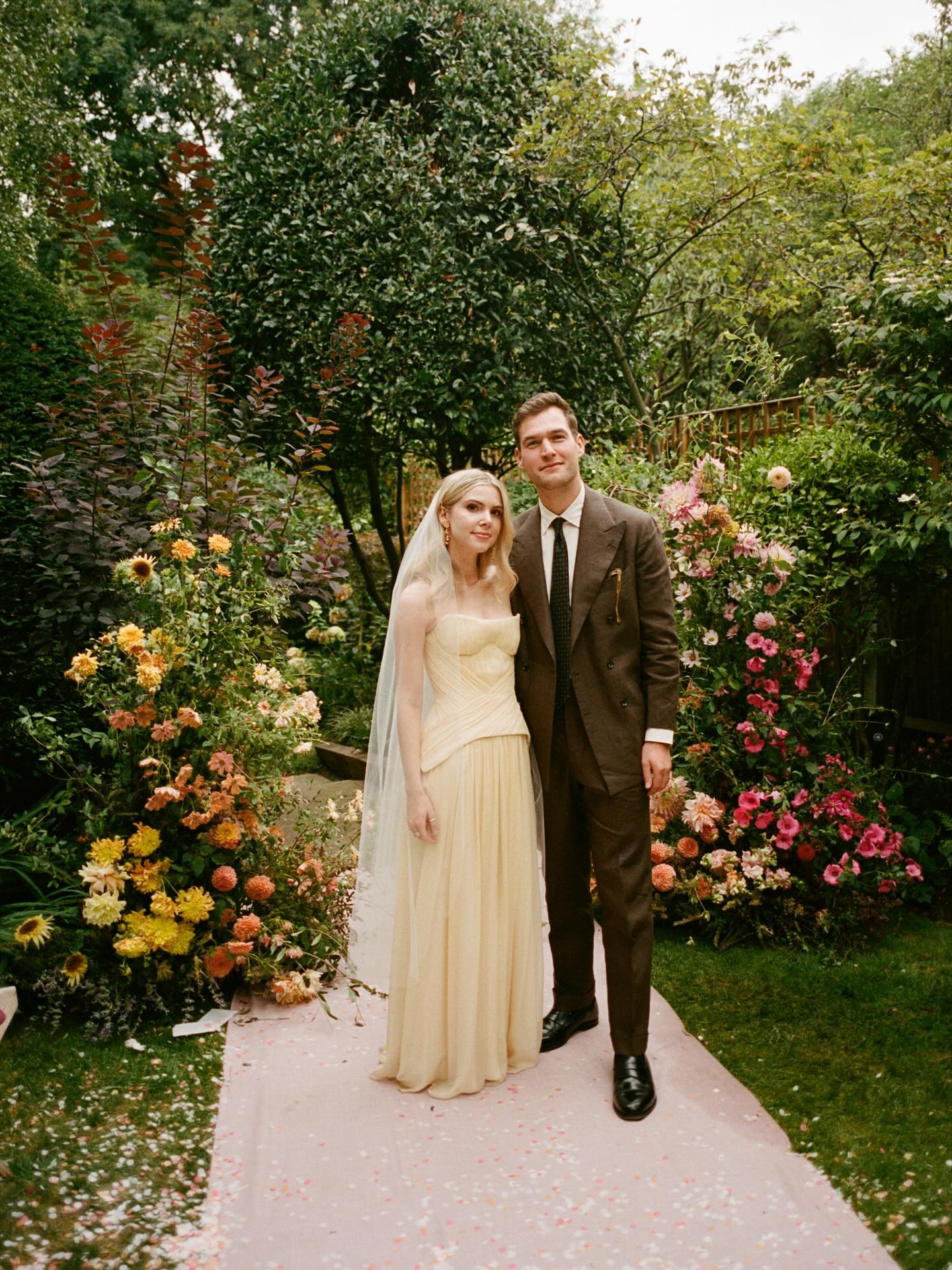 Bride in butter yellow corset wedding gown and veil with groom in chocolate brown suit in a garden filled with pastel florals