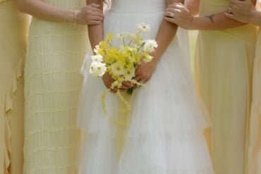 Bride in tiered white tulle wedding gown holding a butter yellow bouquet with bridesmaids in pale yellow dresses