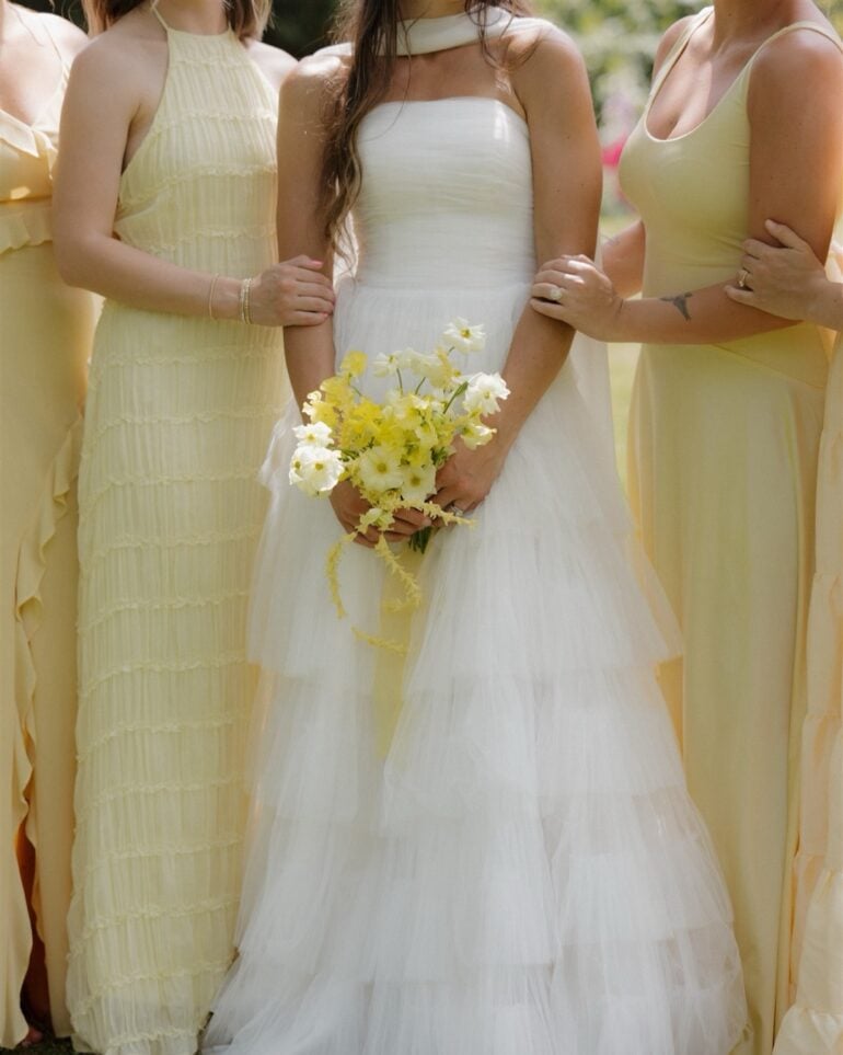 Bride in tiered white tulle wedding gown holding a butter yellow bouquet with bridesmaids in pale yellow dresses